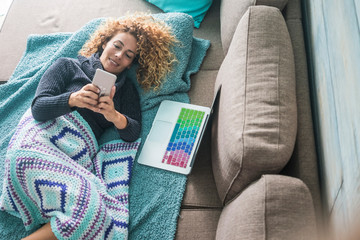 Obraz premium Reaxed caucasian woman type and use mobile phone device lay down on the sofa covered with blue blankets - woman and technology - laptop computer ready for work or onine activity