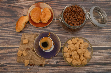 A Cup of coffee, pieces of brown sugar in a sugar bowl, coffee beans in a glass jar and croissants on a wooden background.