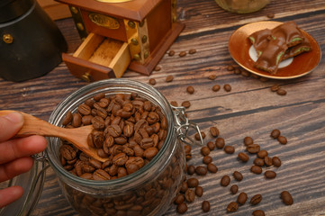 The girl's hand takes a wooden spoon of coffee beans from a glass jar, a coffee grinder, pieces of chocolate on a wooden background. Close up.