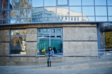 Stylish fashionable african american women in jeans wear and black beret with yellow flowers bouquet posed outdoor in sunny day against blue modern building.