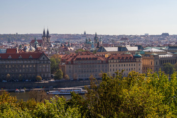 Fototapeta na wymiar City views Prague autumn. Green foliage in the foreground. Tiled roofs. Vlatva river. Bridge. Boats. View from above.