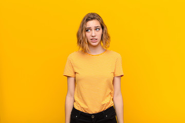 young blonde woman looking worried, stressed, anxious and scared, panicking and clenching teeth against flat color wall