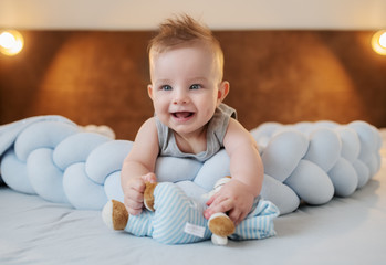 Fototapeta premium Adorable smiling caucasian little baby boy 6 months old lying on stomach on bed in bedroom and holding teddy bear.
