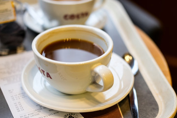 Shallow focus of coffee cup, partially drunk and located on a cafe table. A paper till receipt and another cup is visible.