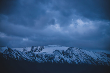 Obraz premium Atmospheric alpine darker landscape to snowy mountain ridge in dusk. Wonderful overcast scenery with snow on mountain peak in dark time. Beautiful snowy top in twilight. Dark violet cloudy sky.