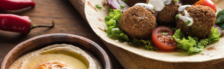 close up view of falafel with vegetables and sauce on pita near hummus on wooden table, panoramic shot