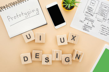 top view of gadgets near wooden blocks with ui and ux design lettering, website design template, notebook with prototyping lettering and green plant on beige background