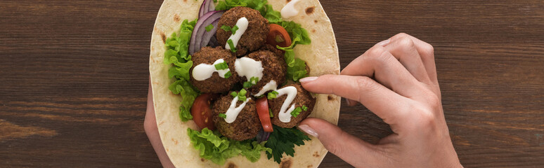 cropped view of woman eating falafel with vegetables and sauce on pita on wooden table, panoramic shot