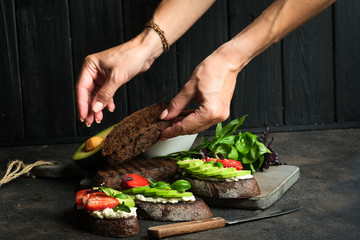 Toast or sandwich with avocado, cheese, strawberries, herbs and seeds on a dark background. Female hands serve a dish. An idea for bruschetta or for a healthy snack. Healthy vegan breakfast.