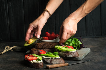 Toast or sandwich with avocado, cheese, strawberries, herbs and seeds on a dark background. Female hands serve a dish. An idea for bruschetta or for a healthy snack. Healthy vegan breakfast.