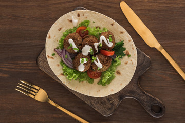 top view of falafel with vegetables and sauce on pita near cutlery on wooden table