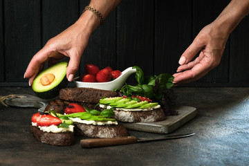 Toast or sandwich with avocado, cheese, strawberries, herbs and seeds on a dark background. Female hands serve a dish. An idea for bruschetta or for a healthy snack. Healthy vegan breakfast.