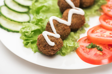 close up view of falafel with sauce on plate with sliced vegetables on white background