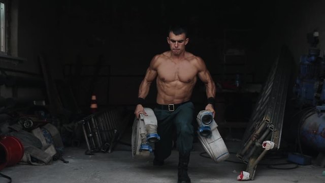 A handsome shirtless firefighter doing lunges holding a firehose in each hand. Fire-fighting equipment on the background