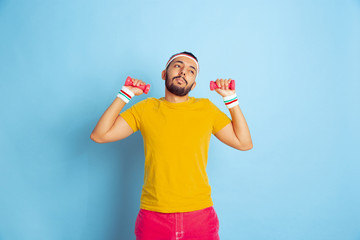 Young caucasian man in bright clothes training on blue background Concept of sport, human emotions, facial expression, healthy lifestyle, youth, sales. Training with the colorful weights. Copyspace.