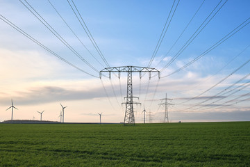 Strommasten aufgereiht bis zum Horizont auf einer grünen Wiese, im Hintergrund Windräder