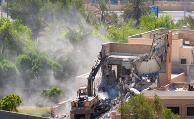 A bulldozer demolishing an old house spreading dust in the air and surrounding trees.
