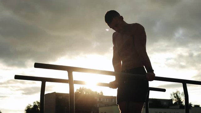 Strong young man doing triceps exercises on parallel bars during street workout. Bottom view. Beautiful sky with the sun and clouds on the background