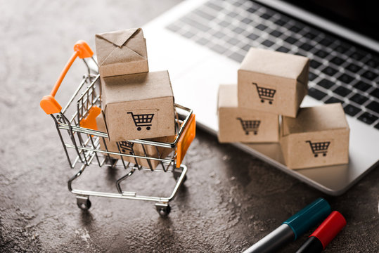 selective focus of toy shopping cart with small carton boxes near laptop, e-commerce concept