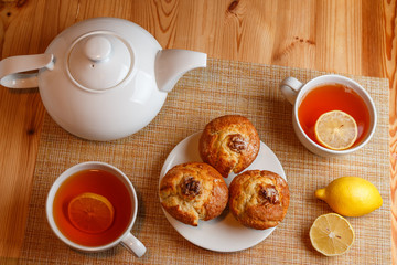 white teapot and two white cups of tea with lemon and muffins top view