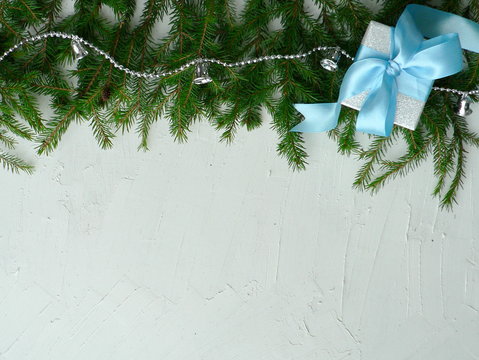 Festive Christmas Border On A White Textured Background With Space For Text. Fir Green Branches Are Decorated With Silver Bells And Beads. Silver Holiday Gift Box With Blue Bow And Ribbon.