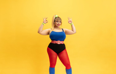 Young caucasian plus size female model's training on yellow background. Stylish woman in bright clothes. Copyspace. Concept of sport, healthy lifestyle, body positive, fashion. Smiling, showing nice.