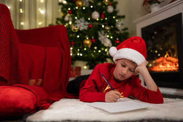 Obraz na płótnie Canvas the boy lies under the Christmas tree near the fireplace and writes a letter to Santa Claus