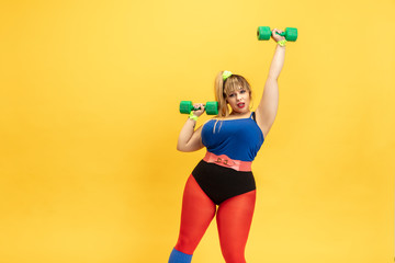 Young caucasian plus size female model's training on yellow background. Stylish woman in bright clothes. Copyspace. Concept of sport, healthy lifestyle, body positive, fashion. Practicing with weights