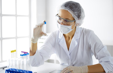 Professional female scientist in protective eyeglasses researching tube with reagents in...