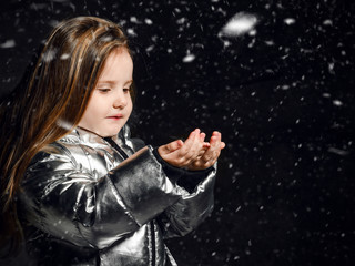 Little girl kid in a winter silver down jacket stands under the snow and catches snowflakes on a...