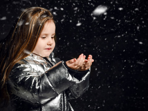 Little girl kid in a winter silver down jacket stands under the snow and catches snowflakes on a dark background