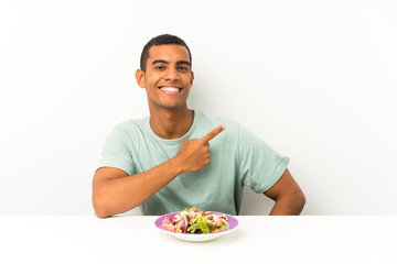 Young handsome man with salad in a table pointing finger to the side