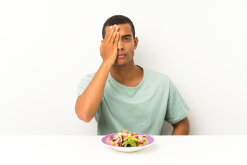 Young handsome man with salad in a table covering a eye by hand