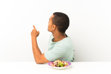 Young handsome man with salad in a table pointing back with the index finger