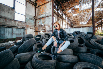 Fototapeta na wymiar Portrait of fashionable blonde girl wearing a rock black style outdoors, sits on old car tires in an abandoned factory