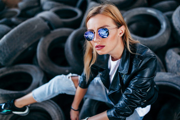 Fototapeta na wymiar Portrait of fashionable blonde girl wearing a rock black style outdoors, sits on old car tires in an abandoned factory