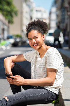 happy young african american girl sitting outside in city holding mobile phone