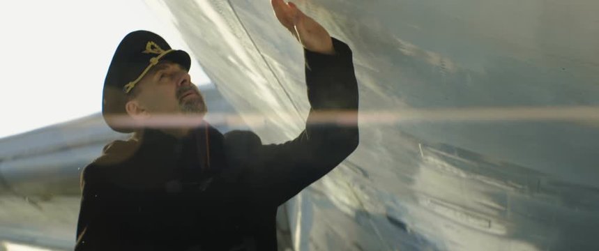 Aged pilot or captain of an aircraft in cap slowly walking on runway under the wing of passenger airplane and touches it. Anamorphic gimbal shot