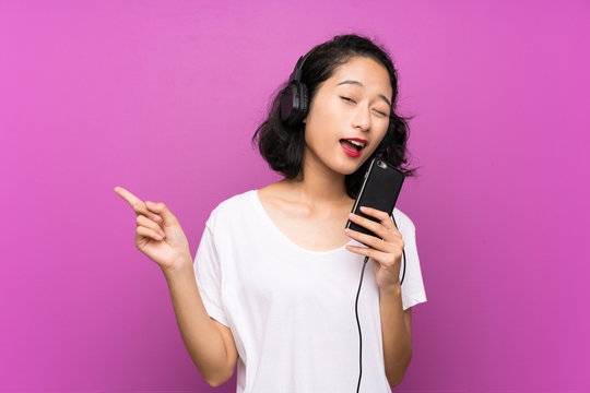 Asian young girl listening music with a mobile and singing over isolated purple wall