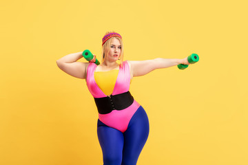 Young caucasian plus size female model's training on yellow background. Copyspace. Concept of sport, healthy lifestyle, body positive, fashion, style. Stylish woman practicing with green weights.