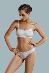 Young woman in white underwear, with bundled hair, hands on hips, posing against gray background....
