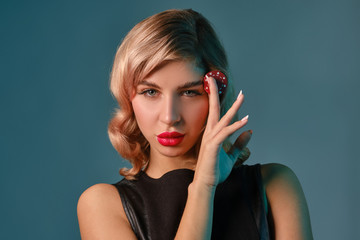 Blonde girl in black leather dress holding one red chip, posing against blue background. Gambling entertainment, poker, casino. Close-up.