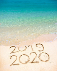 Happy New Year 2020, lettering on the beach with wave and clear blue sea. Numbers 2020 year on the sea shore, message handwritten in the golden sand on beautiful beach background. New Years concept.