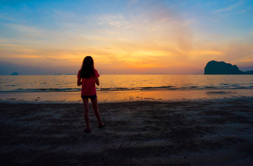 Fototapeta na wymiar Lonely Asian teenager girl standing on the beach watching sunset sky in the evening. Back of a single sad teen standing alone on the beach near the coast of the sea. Pakmeng beach, Trang, Thailand.