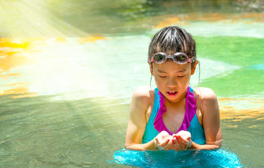 Pretty Asian kid with perfect skin under sun light in swimming pool. Portrait of preteen girl in swimsuit with swimming goggles looking at water in hands in  pool side. Summer holiday vacation concept