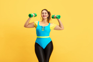 Young caucasian plus size female model's training on yellow background. Copyspace. Concept of sport, healthy lifestyle, body positive, fashion, style. Stylish woman emotional practicing with weights.