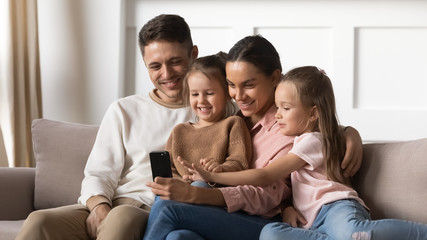 Happy parents with kids daughters relax on sofa using smartphone