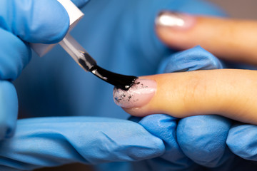 Fingers in blue rubber gloves hold a brush on the client’s fingernail close-up.