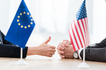 cropped view of diplomat of european union showing thumb up near ambassador of usa sitting with...