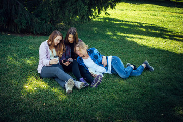 Three young pretty women on the lawn in park look at smartphone. Female students relax in the park sitting on the green grass. Blonde, brunette and brown-haired, top view.